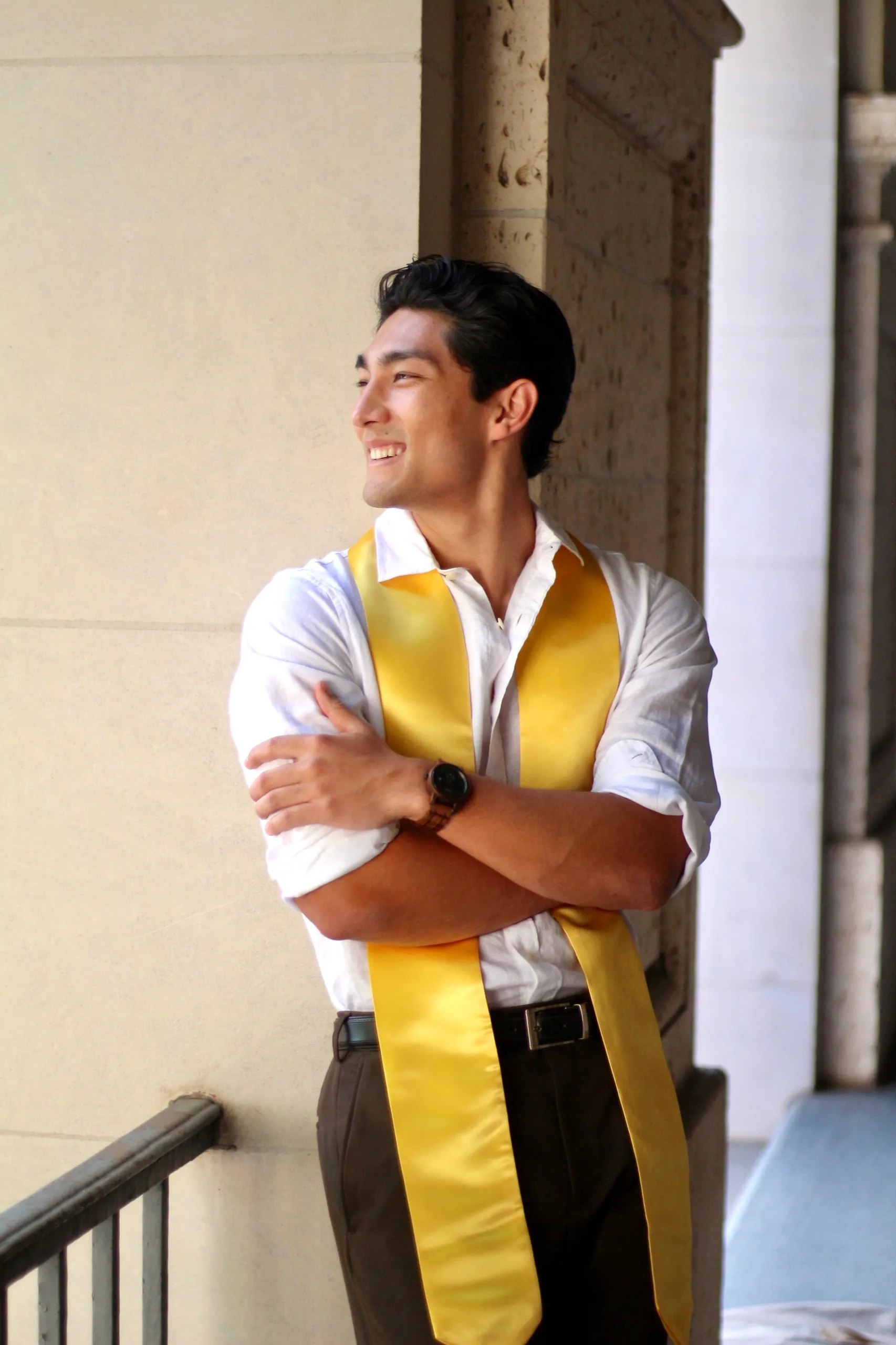 A grad in a white shirt and gold Texas stole smiling with arms crossed beside a stone column.