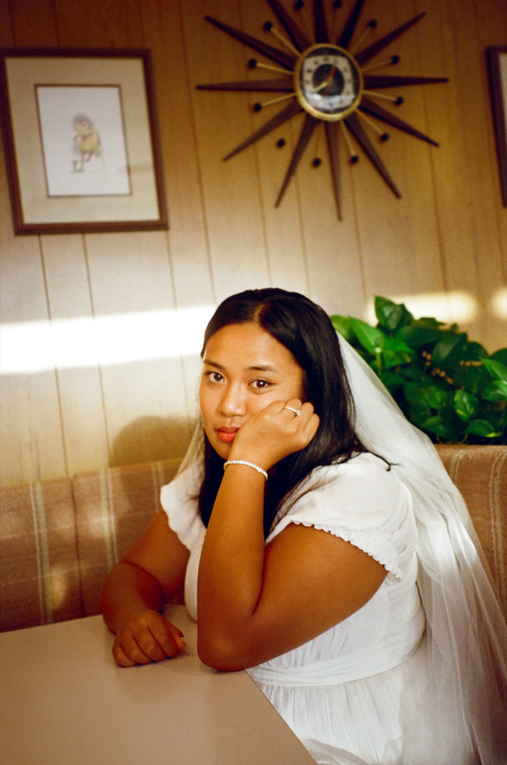 The bride in her veil leaning on a table in a wood-paneled room with a starburst clock on the wall.