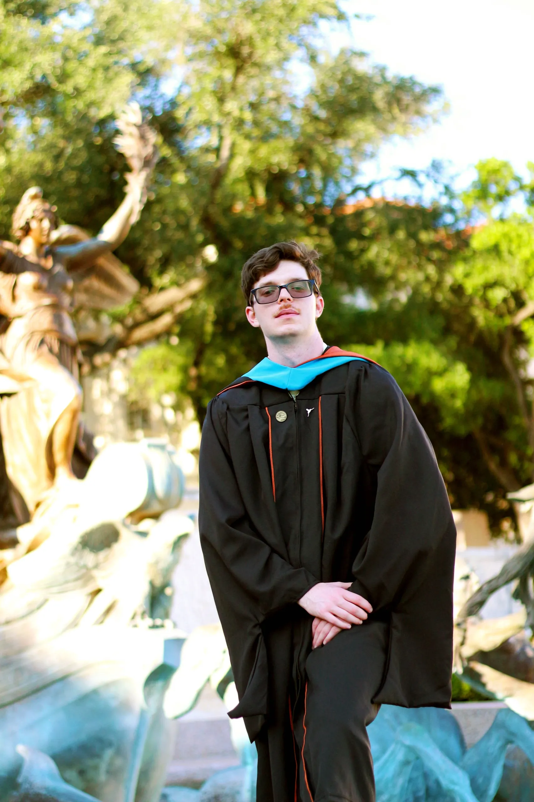 A grad in a black gown with a teal hood standing in front of a bronze fountain sculpture.