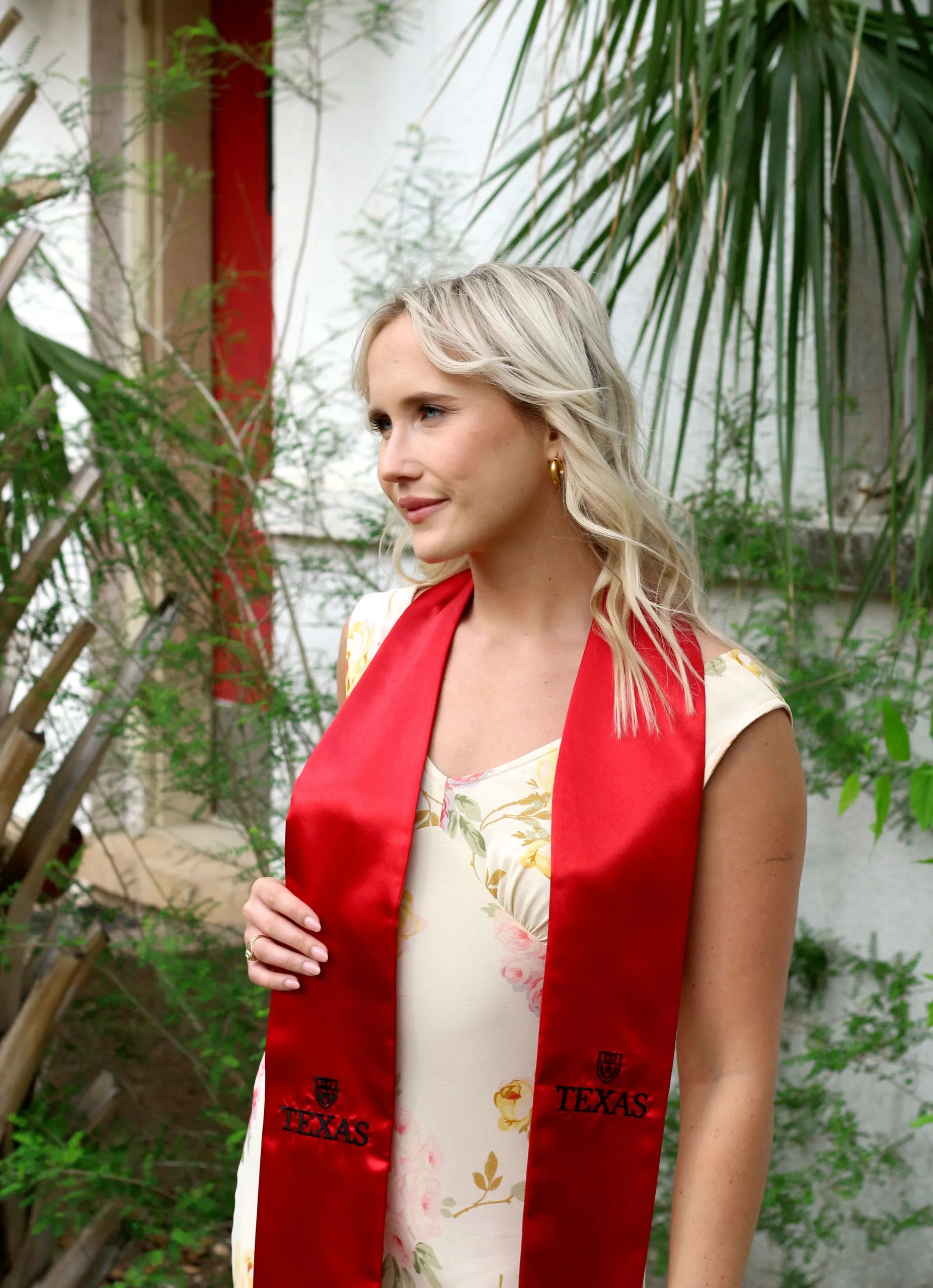 A grad in a floral dress and red Texas stole standing among palm fronds in a garden.