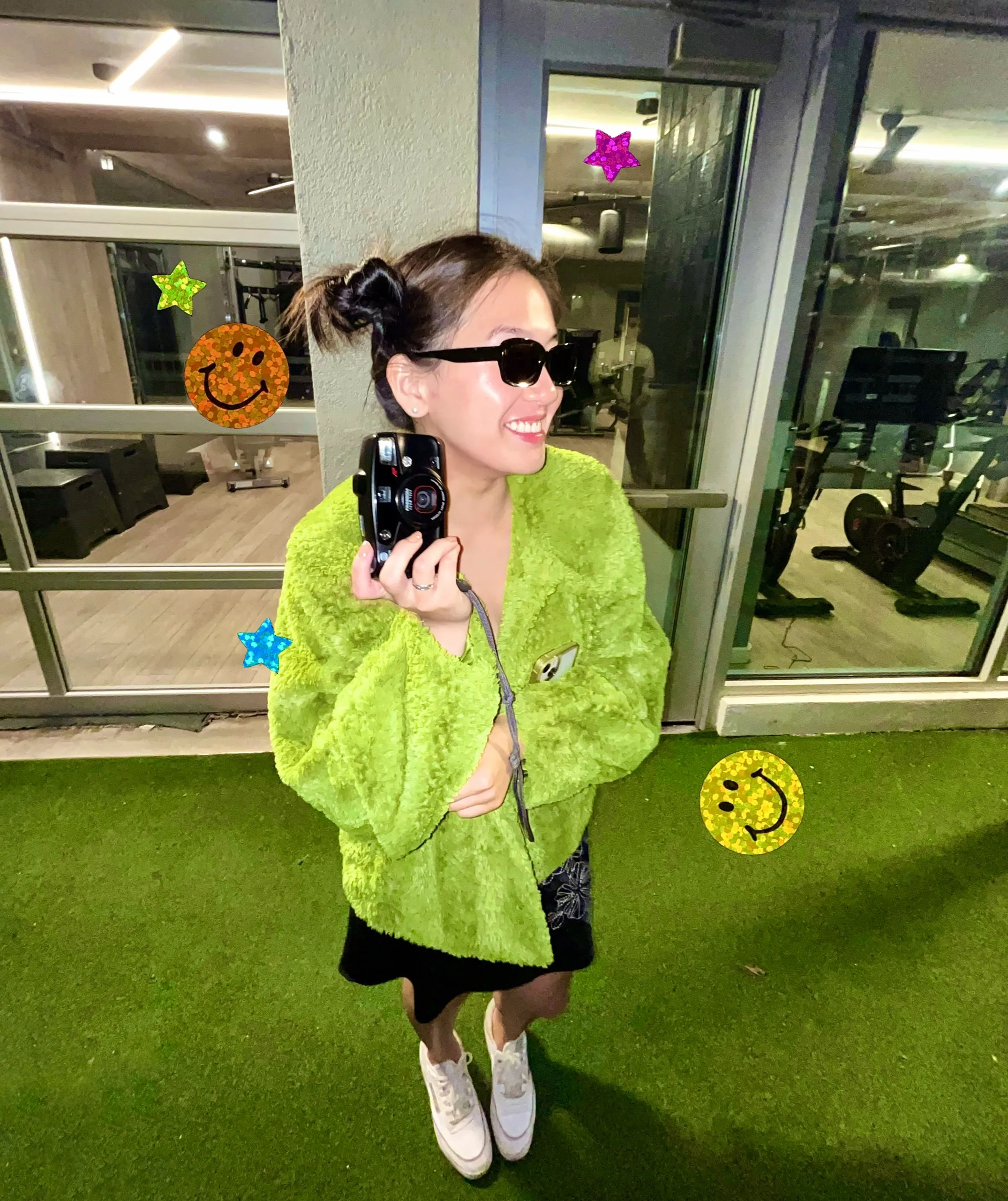 Joie in a fuzzy green coat and sunglasses, holding a camera.