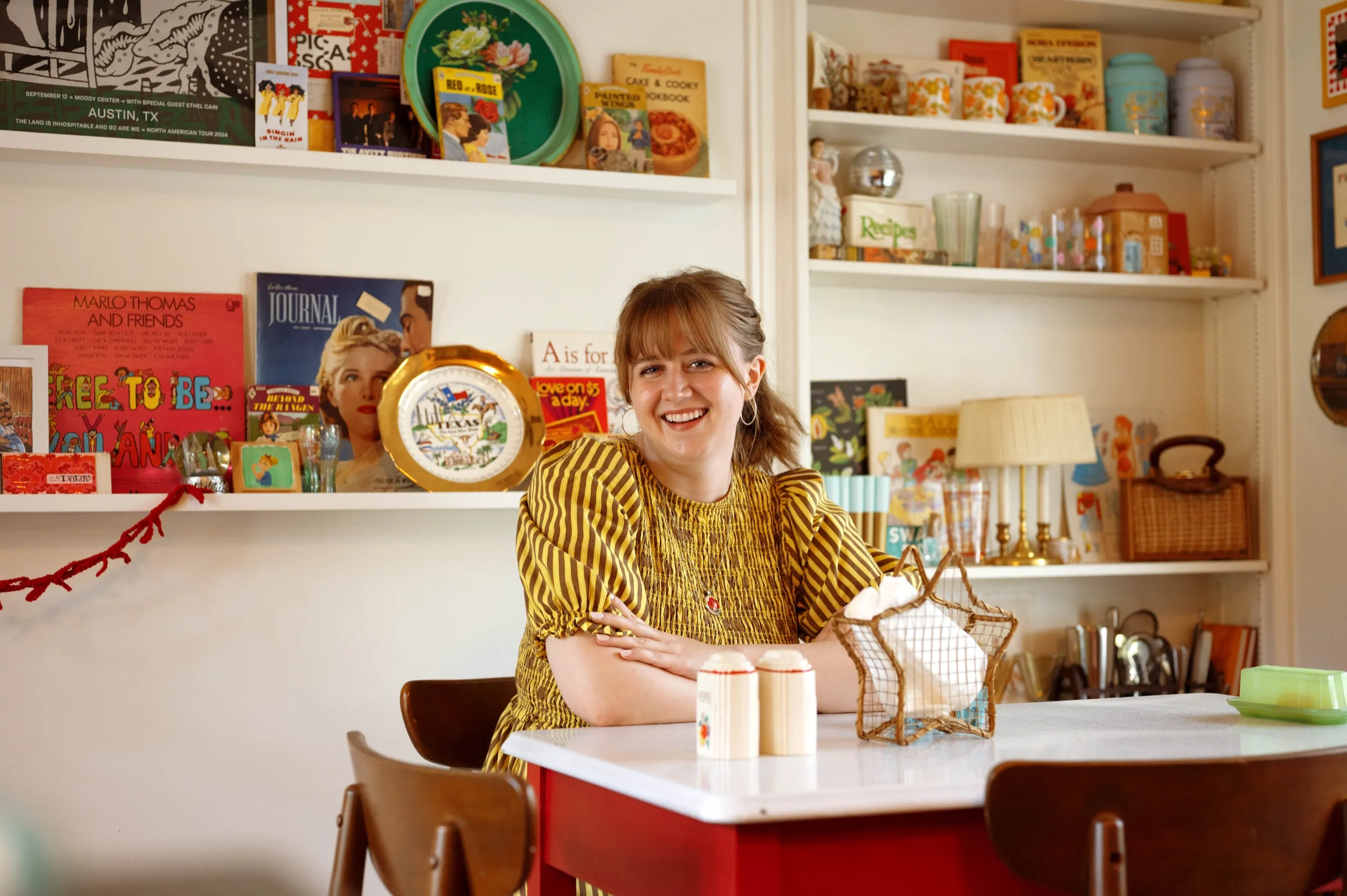 A woman in a striped yellow dress smiling at a red table in a kitchen lined with shelves of vintage tins and books.