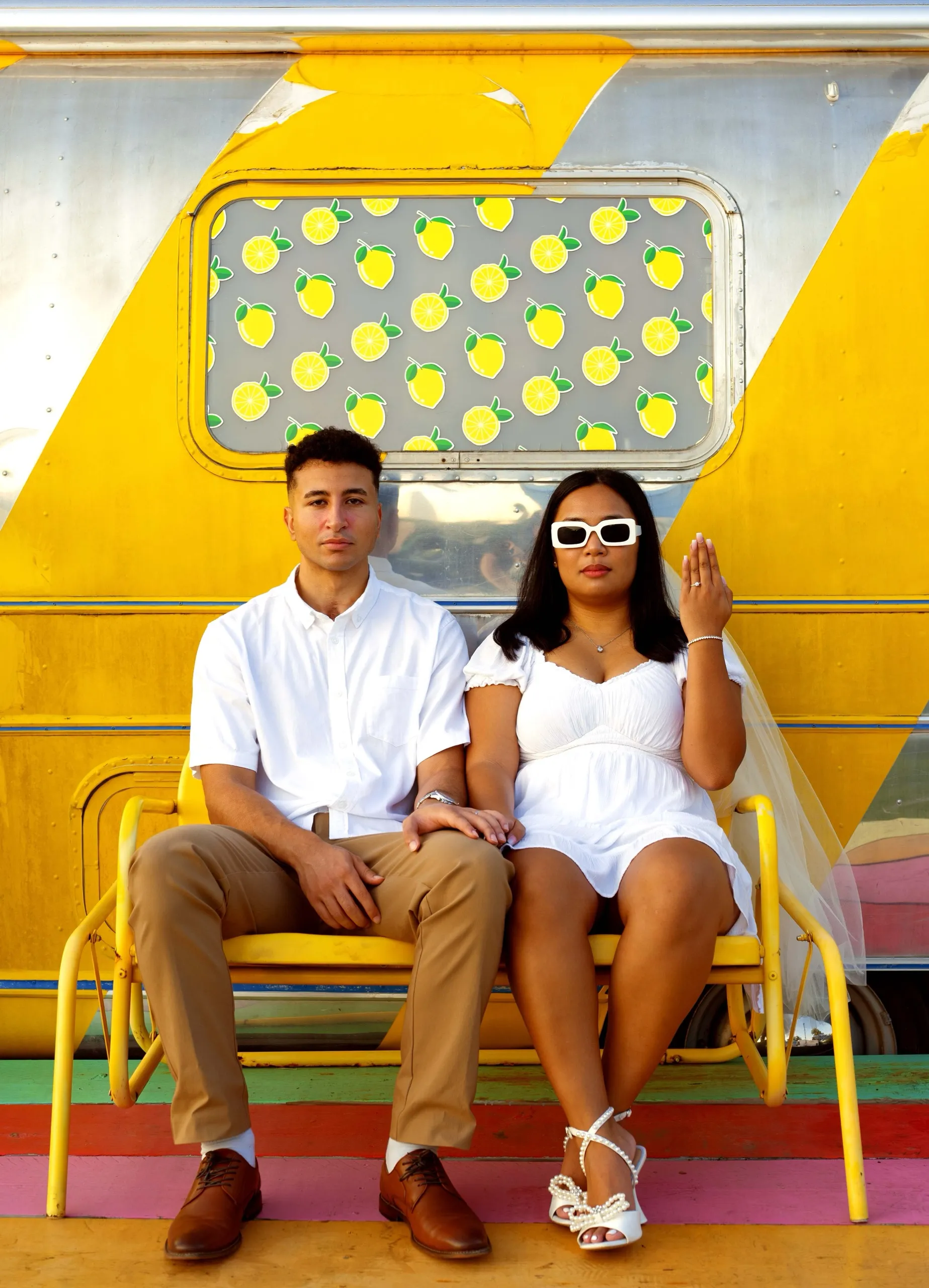 The couple sitting on a yellow bench in front of a silver and yellow trailer painted with lemons.
