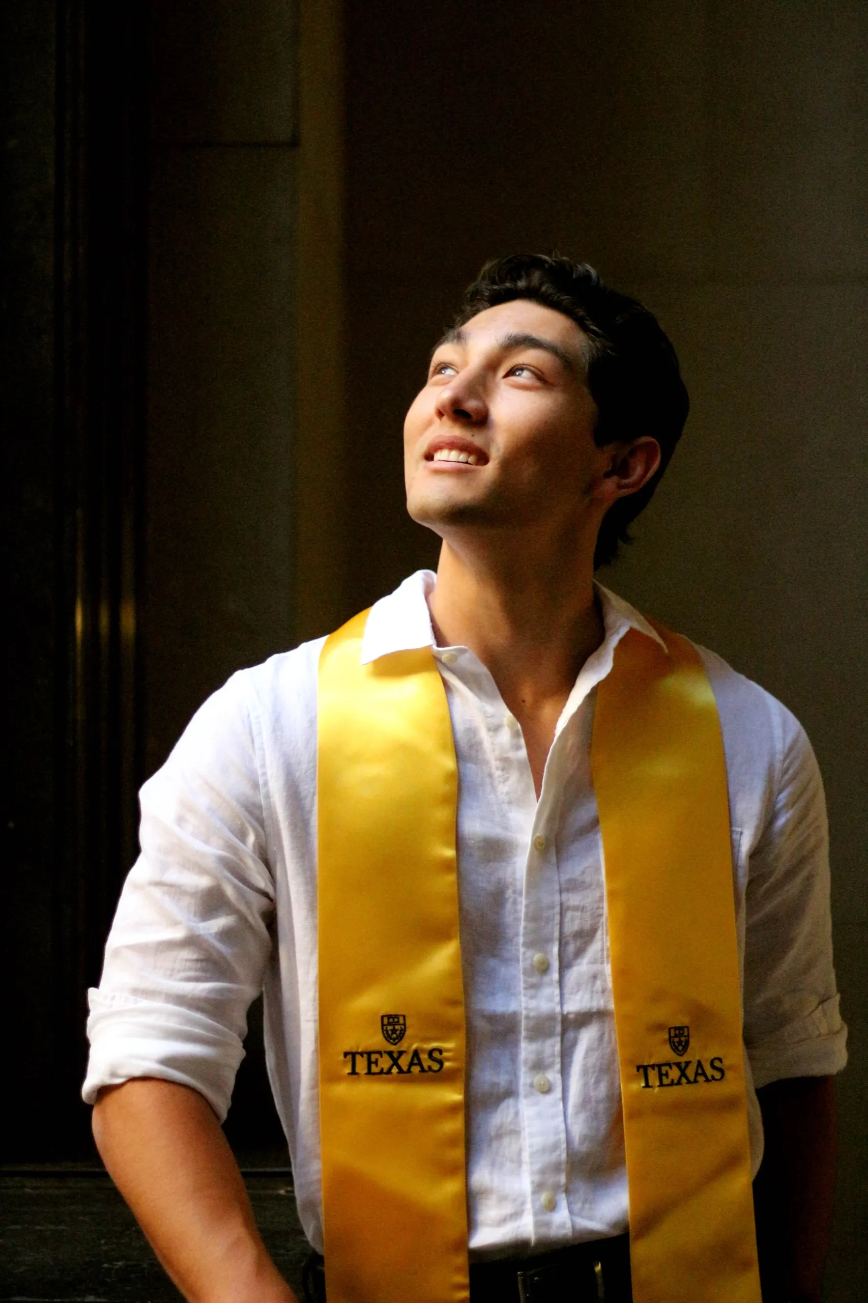 The grad looking up into a beam of light in a dark stone doorway, wearing a gold Texas stole.