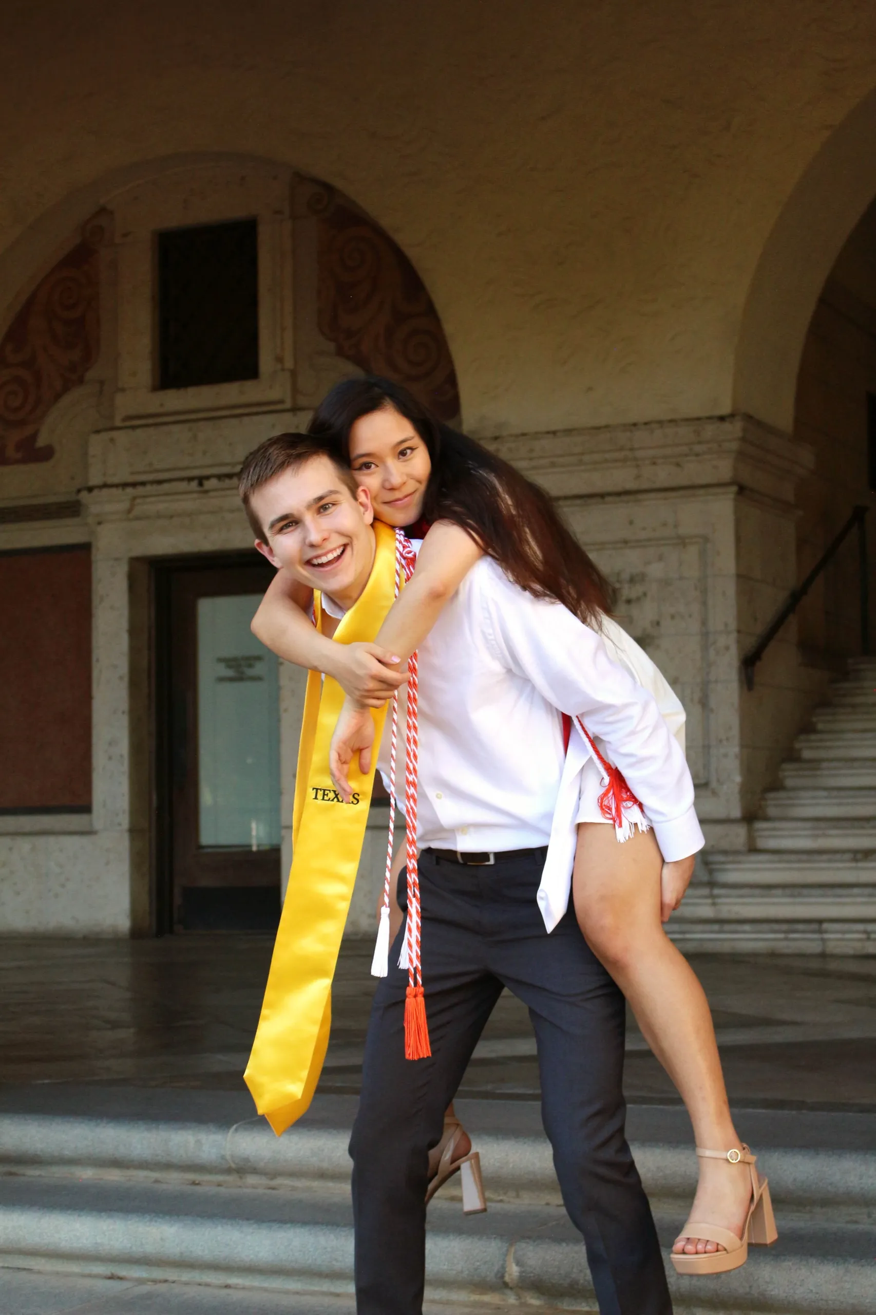A grad in a gold Texas stole carrying their partner piggyback in front of an arched stone building.