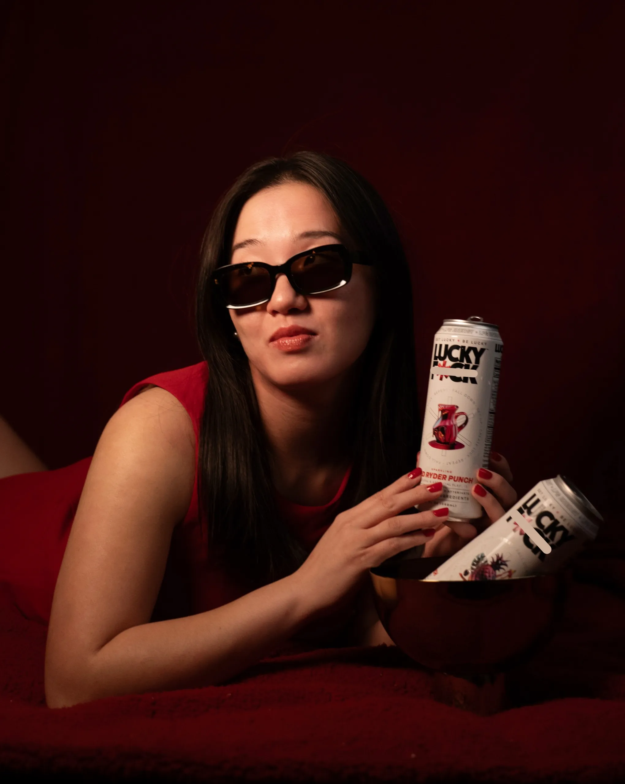 Joie in sunglasses and a red dress, lounging with cans of Lucky energy drink.