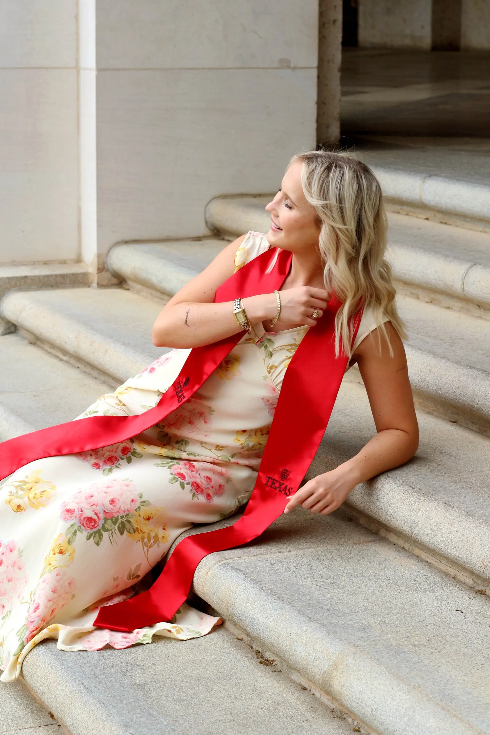 The grad reclining on wide stone steps in a floral dress and red stole.